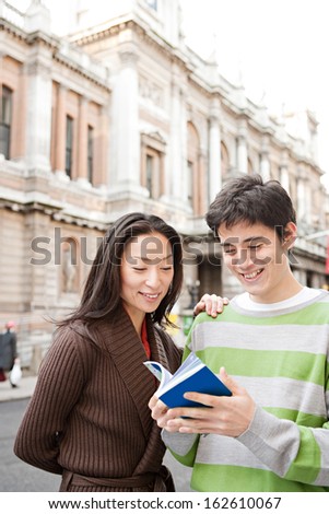 Portrait of a young and attractive Japanese tourist couple visiting the city of London on vacation together and reading a guide book in a classic street, laughing joyfully outdoors.