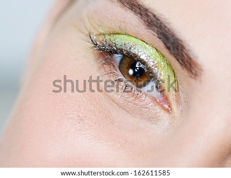 Close up beauty portrait of an attractive woman eye wearing fantasy colorful eyeshadow make up and glitter mascara for a party, smiling and looking at the camera, indoors.