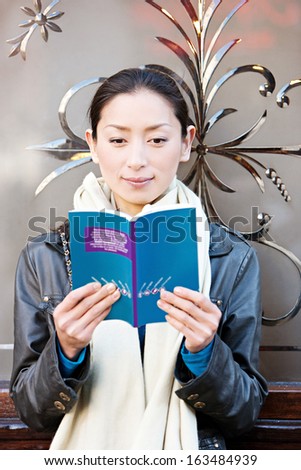 Portrait view of a young attractive Japanese tourist woman in the city of London, reading a travel guide book while leaning on a decorative glass window in an English pub building, outdoors.
