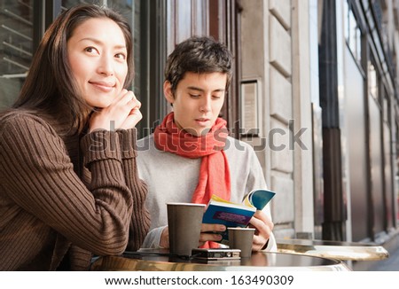 Portrait of a young Japanese couple sitting down at a cafe shop terrace taking a break in the city of London while on vacation, drinking a cup of hot coffee and reading a travel guide book, outdoors.