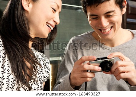 Young and attractive Japanese couple looking at their consumer digital photo camera screen and laughing together during a sunny day outdoors, having fun using technology.