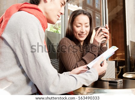 Young and attractive Japanese couple sitting down at a cafe shop terrace in  the city of London while on vacation, drinking a cup of hot coffee and reading a travel guide book, smiling outdoors.