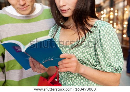 Close up view of a young and attractive Japanese tourist couple in a city shopping mall, holding and reading a travel guide book together, with shops and lights in the background, indoors.