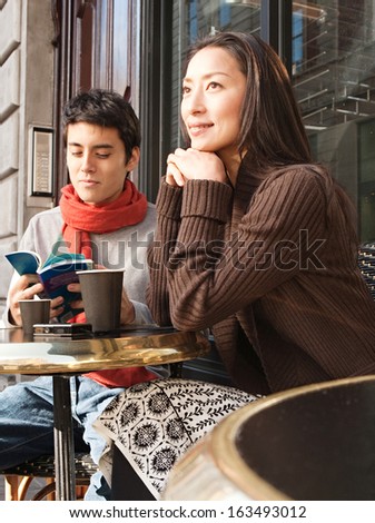Attractive Japanese couple sitting down at a cafe shop terrace taking a break in  the city of London while on vacation, drinking a cup of hot coffee and reading a travel guide book, outdoors.