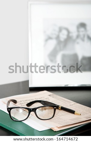 Close up still life of a pair of reading glasses and a black pen laying on an orange financial newspaper on a dark wooden writing desk with a family photo frame. Office interior with no people.