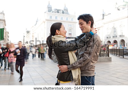 Side portrait view of a young attractive Japanese tourist couple in Piccadilly Circus landmark street hugging and being romantic while visiting the city of London on vacation, smiling outdoors.