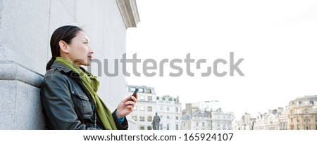 Side view of an attractive Japanese tourist woman relaxing in Traffalgar Square landmark in the city of London, holding a smart mobile phone and relaxing during a sunny day on holiday.