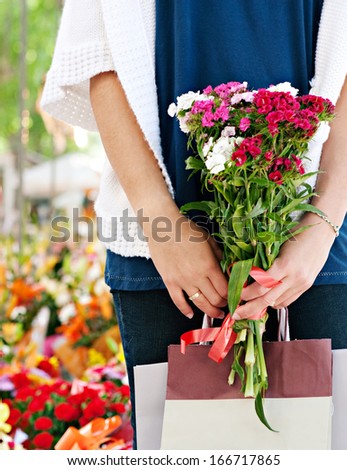 Close up of the middle section of a woman body standing in a fresh flowers market with shopping paper bags and holding a bouquet of flowers in her hands, detail faceless view.
