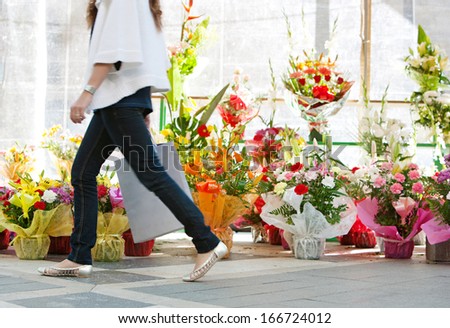 Side view of the lower body section of a young woman walking passed a fresh flowers market stall carrying a paper shopping bag during a sunny day outdoors. Faceless figure.