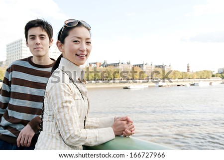 Portrait view of an attractive Japanese tourist couple visiting the city of London on holiday and leaning on the banister of a bridge while contemplating the river Thames during a sunny day outdoors.