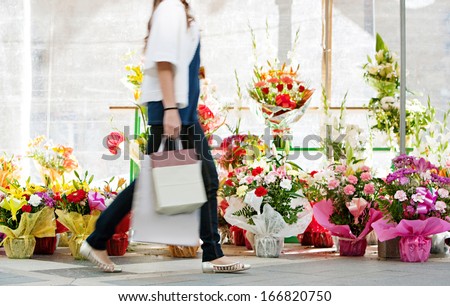 Side view of the lower body section of a young woman walking passed a fresh flowers market stall carrying a paper shopping bag during a sunny day outdoors. Faceless figure.