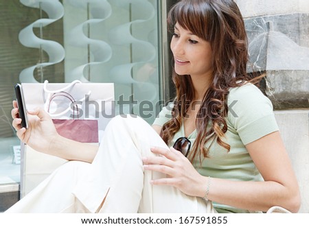 Close up side view of an attractive young woman sitting on the steps of a store window display, smiling, holding and using her smartphone during a shopping day out, outdoors.