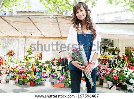 Beautiful young woman buying and holding a small bouquet of fresh flowers from a city floral market stall with colorful arrangements of fresh flowers, during a sunny day outdoors.