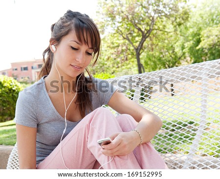 Sporty young woman taking a break from exercising and sitting on a bench, holding a smartphone and listening to music during a sunny day in the park, relaxing outdoors.