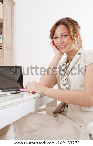 Side portrait view of a young professional business woman using a laptop computer while sitting at her work desk and smiling at the camera leaning on her hand. Office interior.