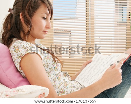 Side view of a student woman studying and holding a dictionary open whilst reading it in preparation for her college exams. Home interior bedroom.