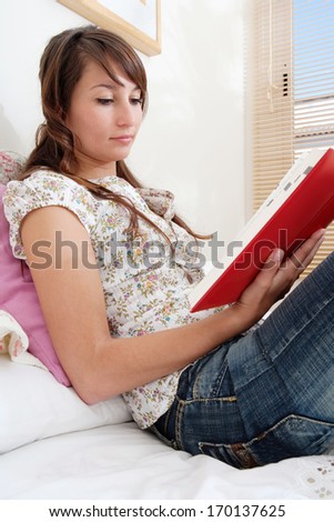 Side view of a student woman studying and holding a dictionary open whilst reading it in preparation for her college exams. Home interior bedroom.
