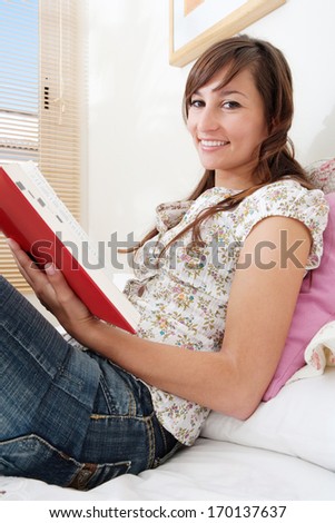 Side view of a student woman studying and holding a dictionary open whilst reading it in preparation for her college exams, smiling at the camera. Home interior bedroom.