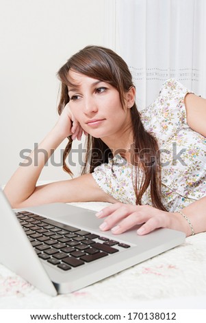 Attractive young woman laying down on her bed at home using a laptop computer and browsing on line. Searching, reading and studying in a home interior.