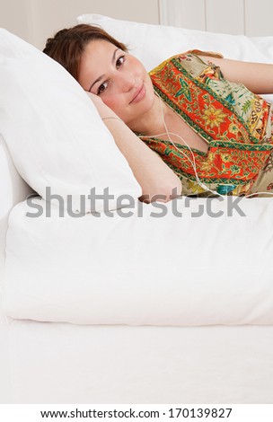 Portrait view of a beautiful young woman laying down on a white sofa in a living room at home, listening to music with her small headphones, being tranquil and relaxing. Home interior.