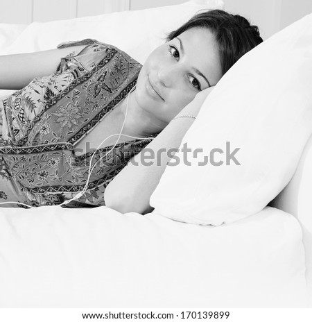 Black and white portrait of a beautiful young woman laying down on a white sofa in a living room at home, listening to music with her small headphones, being tranquil and relaxing. Home interior.