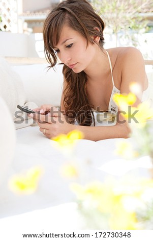 Close up portrait of an attractive young woman laying down and relaxing on a garden outdoors bed at home and using a smart phone during a sunny summer day. Home technology lifestyle.