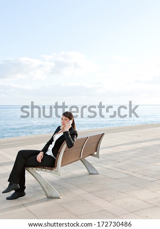 Businesswoman sitting down on a bench on a walk by the sea, relaxing and having a mobile phone conversation using her smartphone during a sunny day at work. Business person outdoors.