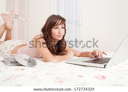 Portrait of a beautiful young woman relaxing on her bed at home using her laptop computer and typing, with films and music DVD\'s. Home entertainment interior lifestyle technology.