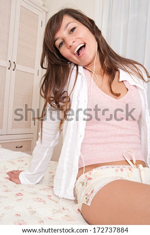 Portrait of an attractive young woman relaxing in her home bedroom and enjoying listening to music using her headphones and singing aloud, smiling at the camera. Home interior technology.