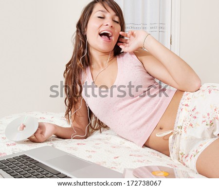 Beautiful young woman relaxing on her bed at home while listening to music with her laptop computer and headphones, singing joyfully. Home interior lifestyle technology.