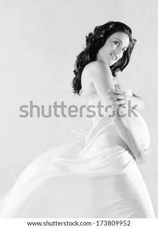Black and white profile portrait of an attractive pregnant woman holding her belly with her hand while wearing a floating silk fabric dress against a plain background. Interior pregnancy beauty care.