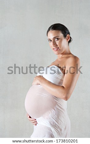 Side portrait view of an attractive pregnant woman holding her belly with her arms while wearing a wet white cotton dress around her body. Interior pregnancy beauty care.