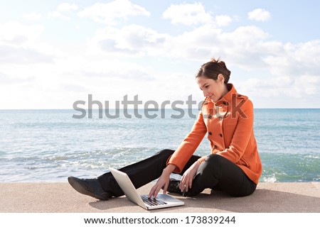 Profile view of a young professional woman sitting down by the ocean with a sunny blue sky, using a laptop computer and working on it during a break. Outdoors technology.