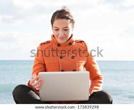 Frontal view of a young professional woman sitting down with her legs crossed by the ocean with a sunny blue sky, using a laptop computer and working on it during a break. Outdoors technology.