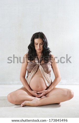 Attractive pregnant woman sitting down on a yoga position, meditating and relaxing while wrapped in transparent silk fabric against a spacious plain background. Interior pregnancy health and beauty.