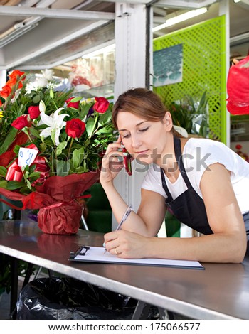 Portrait of an attractive shop assistant woman and small business owner using her smartphone to take an order over the phone in her florist kiosk market stall store. Outdoors business.