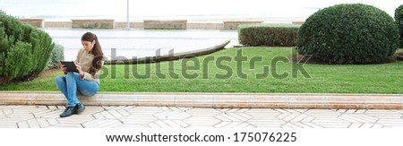 Panoramic view of a park by the sea with a young attractive woman sitting down on a stone step by the grass, using her tablet technology to go on-line and use the internet. Outdoors lifestyle.