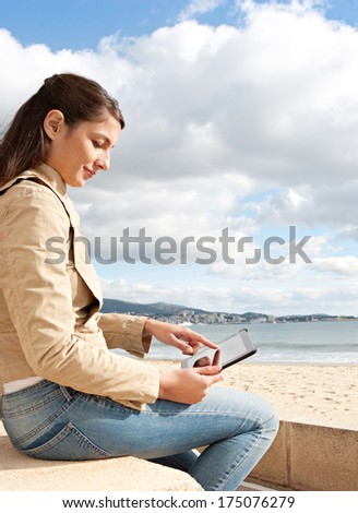 Side view of an attractive young woman sitting on a stone bench by the ocean sea using a digital tablet with touch screen during a sunny day out. Outdoors technology lifestyle.