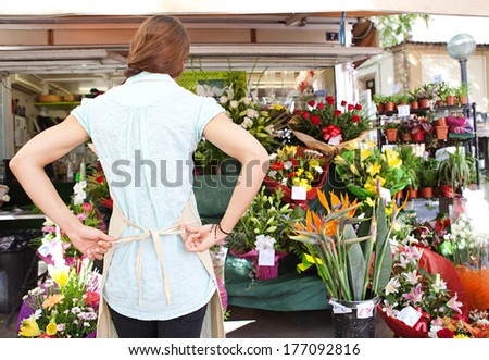 Rear view of a young florist businesswoman fastening her working apron getting ready for the day work and opening a fresh florist store in a flower market, outdoors business life.
