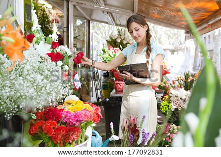 Side view of a florist small business owner checking her fresh flowers stock and inventory holding a clipboard, managing a market stall on a sunny day, outdoors. Running a small business.