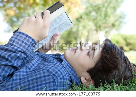 Close up profile portrait of a student teenager boy laying down and relaxing on green grass in a park, reading a text book and studying during a sunny day. Outdoors lifestyle.