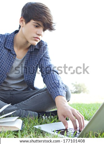Attractive teenager boy sitting down on green grass in a park reading an open reference book and using a laptop while studying and doing his homework against a sunny sky. Outdoors lifestyle.