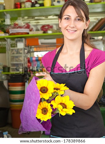 Attractive florist business owner woman working at the counter of her flower market store wrapping yellow sunflowers in wrapping paper during a sunny day. Small business owner.
