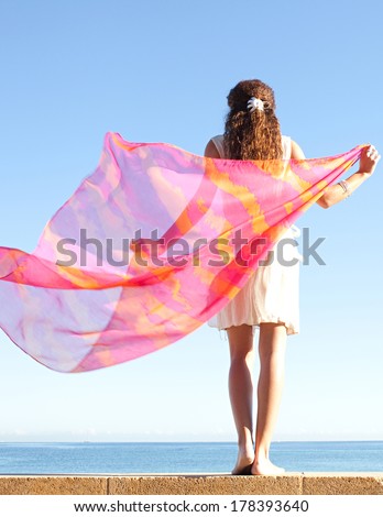 Rear view of a young woman rising a bright and colorful floating sarong fabric up with her arms against a bright blue sky and sea on a holiday beach, outdoors. Travel and lifestyle.