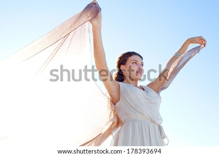 Beauty portrait close up of an elegant thoughtful woman rising a floating silk fabric up with her arms against a bright blue sky on holiday, outdoors. Travel, beauty and lifestyle.