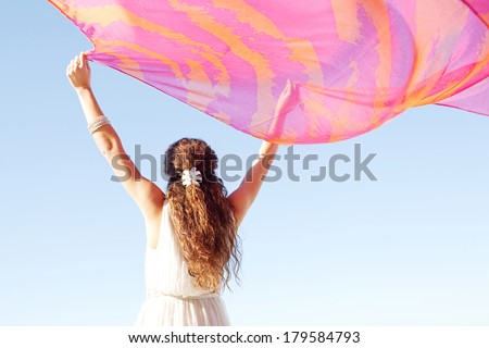 Rear view of a young woman with her arms up in the air holding a pink silk fabric floating with the breeze during a sunny summer day on holiday, against a bright blue sky. Outdoors lifestyle.