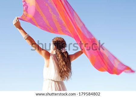 Rear view of a young woman with her arms up in the air holding a pink silk fabric floating with the breeze during a sunny summer day on holiday, against a bright blue sky. Outdoors beauty lifestyle.