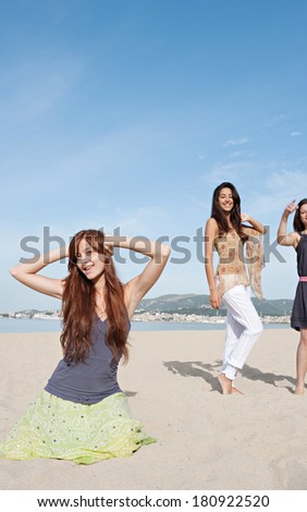 Group of three teenagers female friends enjoying a day on the beach together, listening to music and dancing, celebrating the summer holidays during a sunny day with a blue sky.