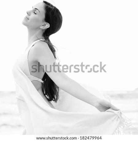 Close up black and white side portrait of a beautiful woman relaxing on a beach on holiday holding a pink fabric sarong around her body and floating in the breeze, breathing fresh air, lifestyle.