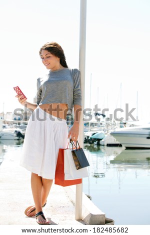 Beautiful young woman relaxing in a boats and yachts marine port carrying paper shopping bags and using a smartphone during a sunny day on holiday. Travel technology lifestyle.